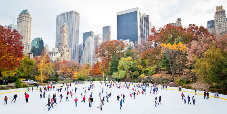 People are ice skating on an outdoor rink in a park surrounded by autumn trees and tall city buildings, one of the classic things to do in New York City in winter under a partly cloudy sky.