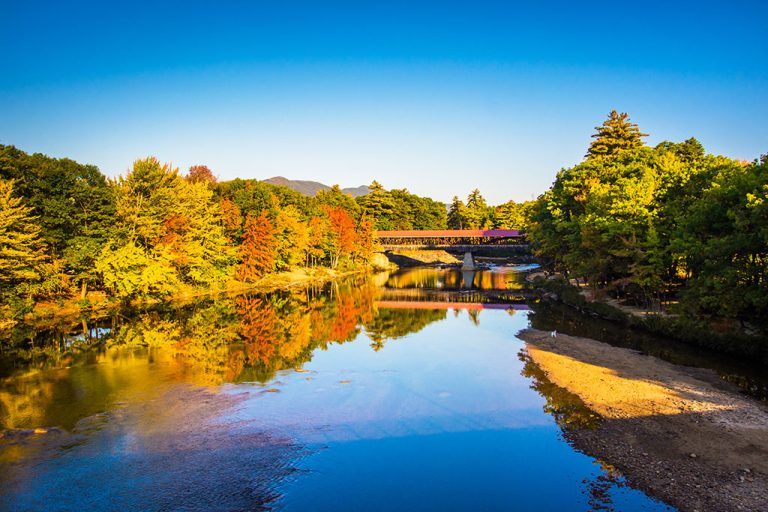 A river reflecting colorful autumn trees and a covered wooden bridge—a scene that captures New Hampshire's fame—crosses the water under a clear blue sky, with sand and rocks lining the riverbank.