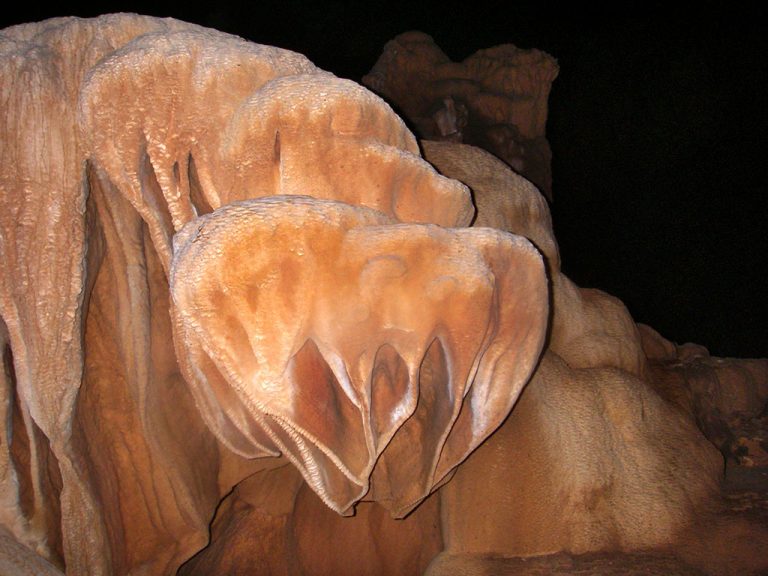 Large brown rock formation with smooth, undulating surfaces and layered, curtain-like structures, illuminated against a dark background. The surface appears textured and slightly shiny, reminiscent of the famous ATM Cave in Belize.
