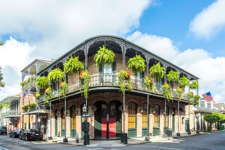 A two-story brick building on a corner with wrought-iron balconies, hanging plants and ferns, wooden shutters, and an American flag—a scene that captures what Louisiana is known for: charming architecture and vibrant Southern character.