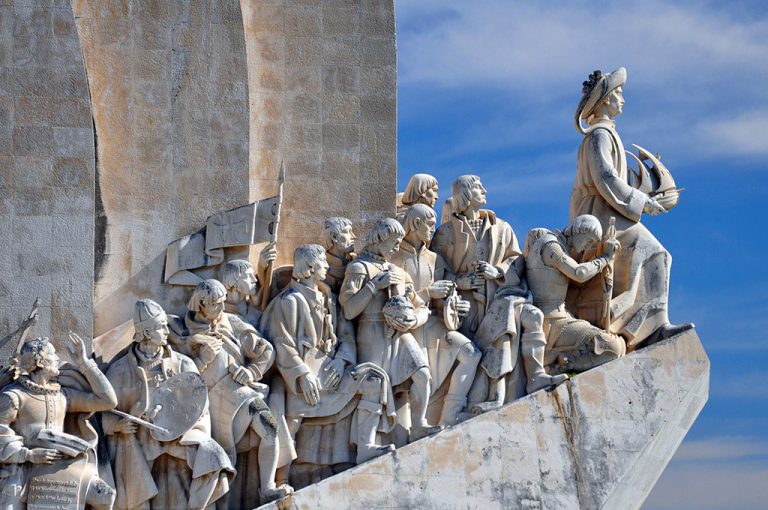 A stone monument features a group of detailed statues, mostly men in historical costume, standing on a sloped surface, with one at the front holding a model ship—an iconic scene that highlights what Lisbon is known for against a vibrant blue sky.