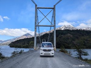 Antes de viajar ao Chile, imagine uma van branca viajando por uma ponte suspensa, emoldurada por montanhas cobertas de neve e florestas exuberantes. O céu está parcialmente nublado enquanto esta rota cênica se estende sobre águas brilhantes, oferecendo um vislumbre das paisagens cativantes do Chile.