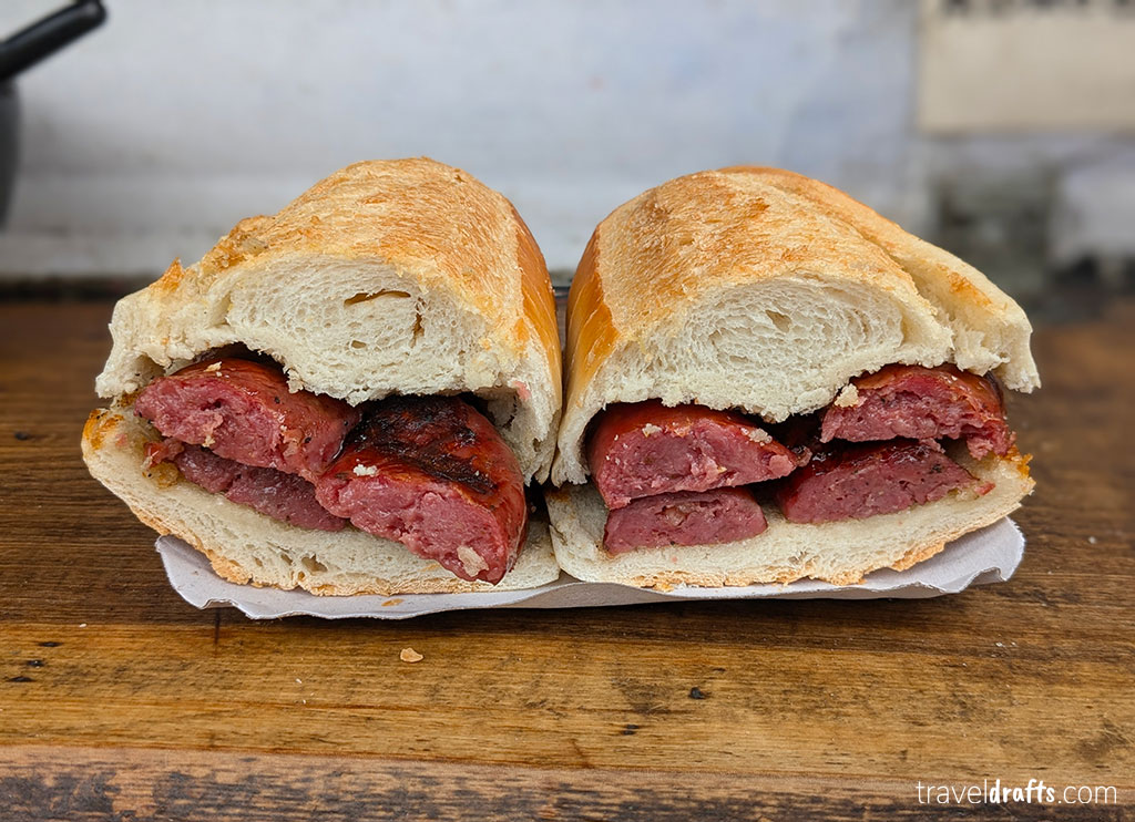 A close-up of a halved sandwich with thick grilled sausages inside a crusty baguette—an irresistible example of Argentine food—displayed on white paper on a wooden surface.