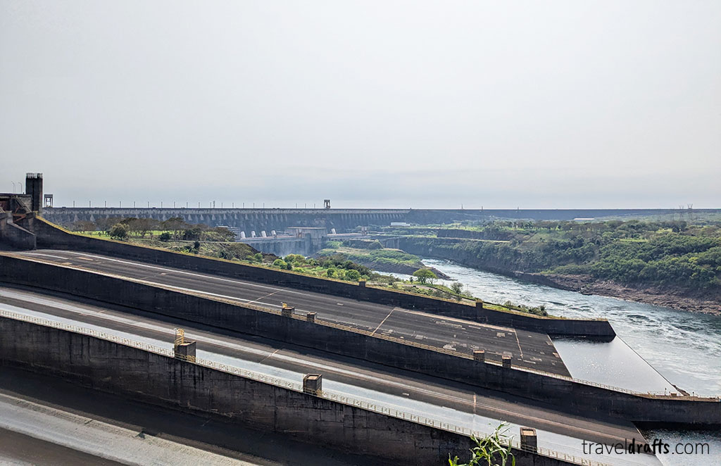 A large dam structure spans a wide river, with concrete channels and spillways in the foreground. Water flows over the spillways, and lush vegetation lines the riverbanks—a stunning sight for anyone visiting Paraguay.