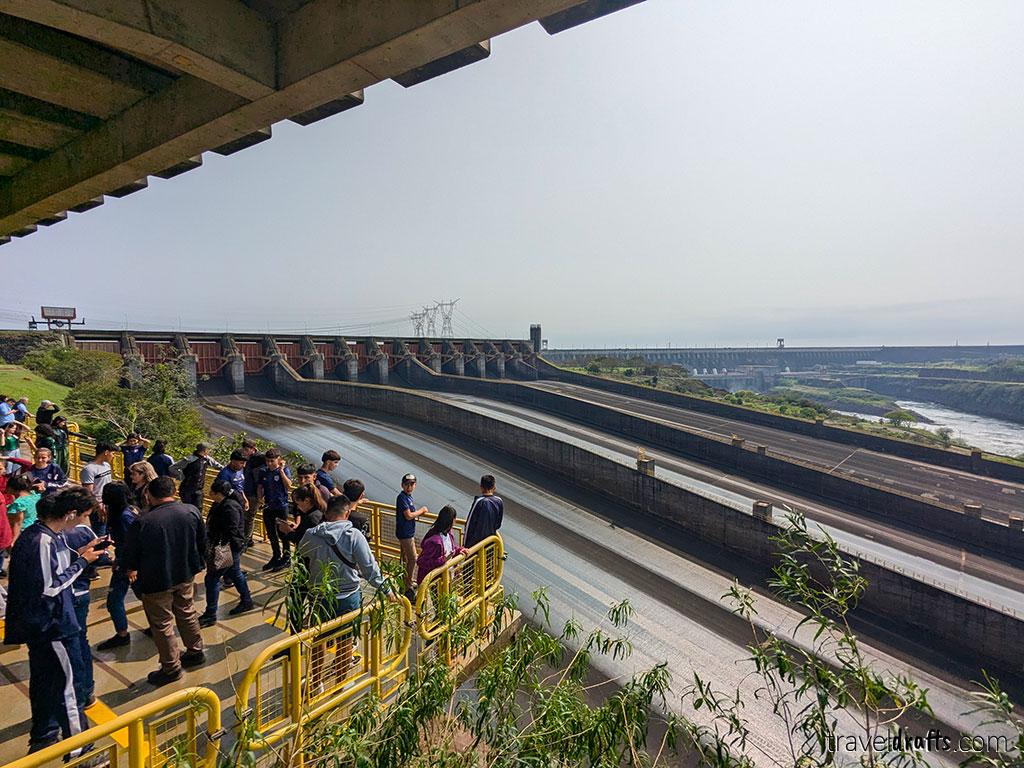 A group of people stand on a viewing platform with yellow trails overlooking a vast dam structure in Paraguay, with water channels below and power lines in the distance—a perfect spot for anyone looking for Paraguay tips on their trip.