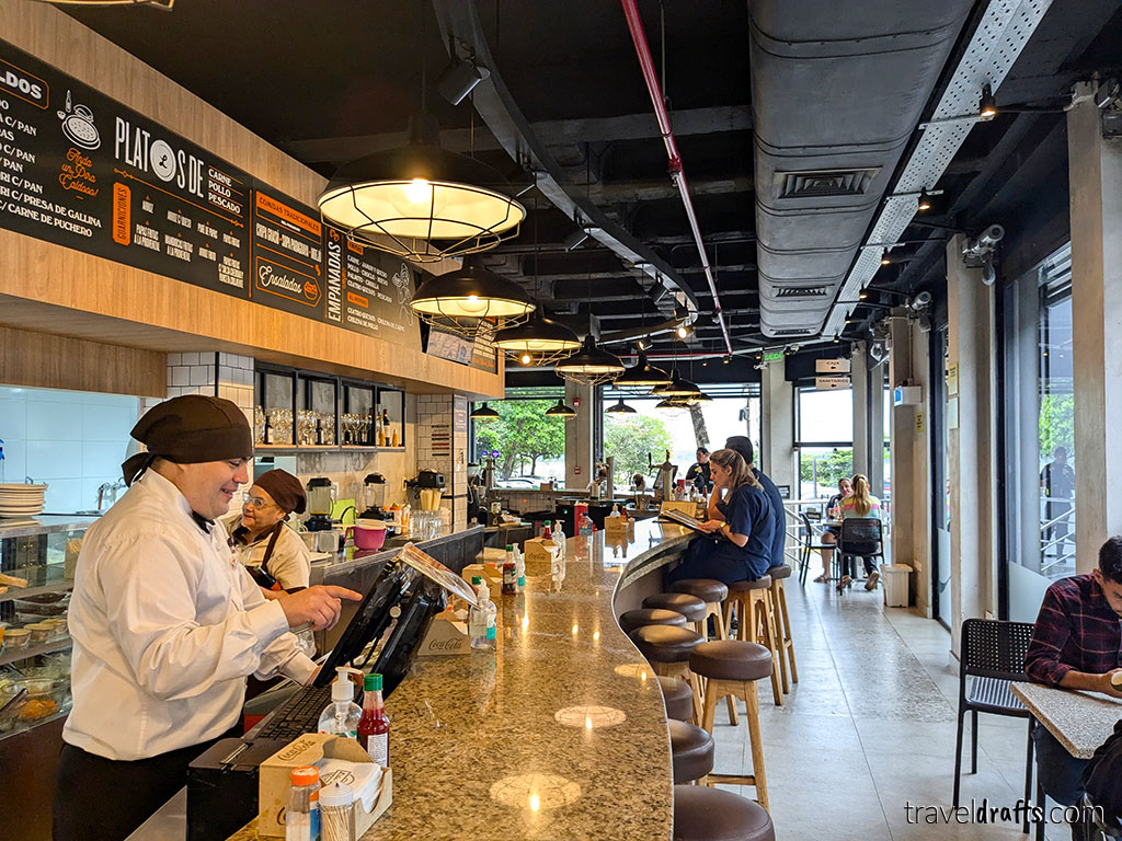 A barista in a white shirt and black cap stands behind a marble counter in a modern café—an ideal spot to relax after traveling through Paraguay. Customers dine and work on high stools, beneath hanging lights and exposed pipes in the ceiling.