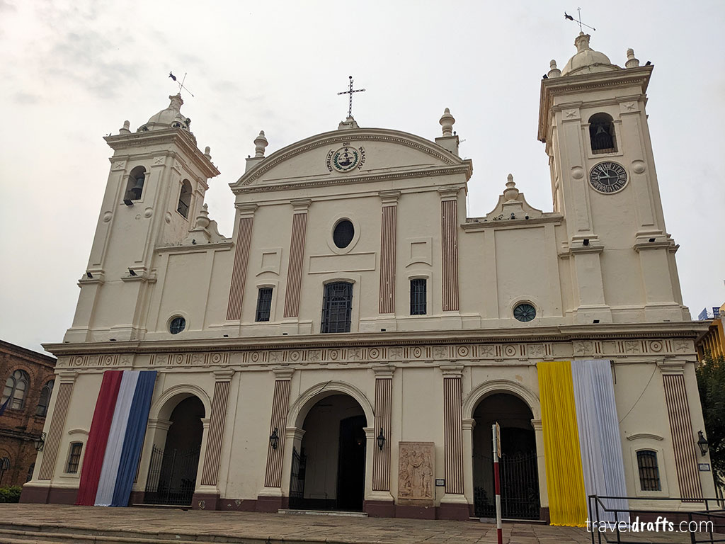 A large cream-colored church with two bell towers, three front entrances, and columns. Vertical red, blue, white, and yellow banners hang on either side—a must-see if you plan to visit Paraguay. The sky is cloudy.