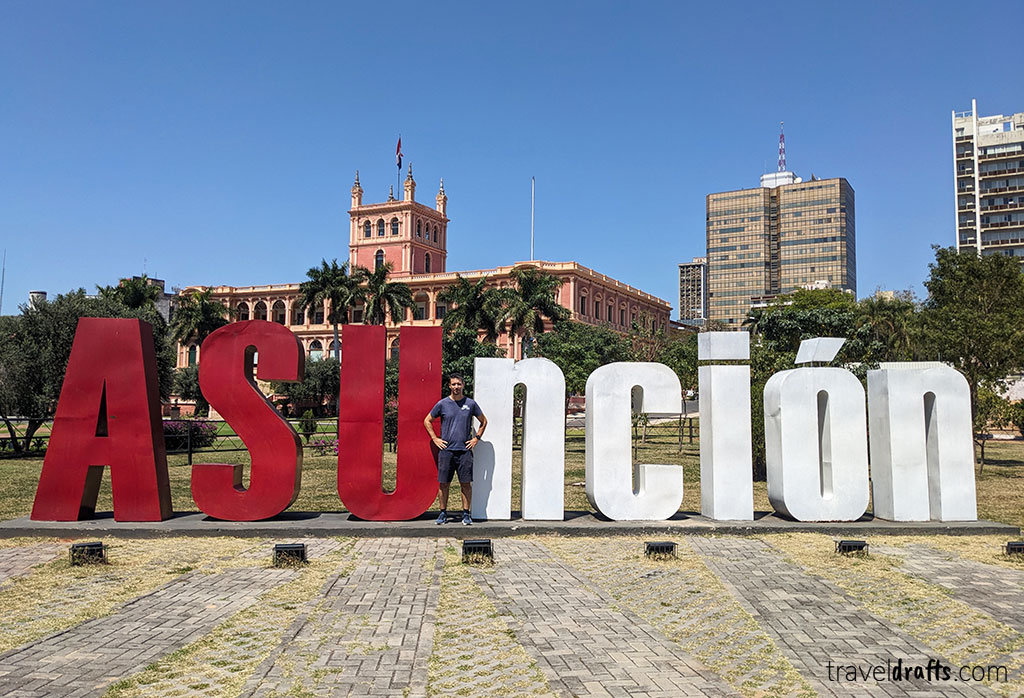 A person stands amid large red and white letters spelling "Asunción" in an open-air plaza, perfect for those looking for tips on places to visit in Paraguay, with a historic pink building and tall, modern buildings in the background.
