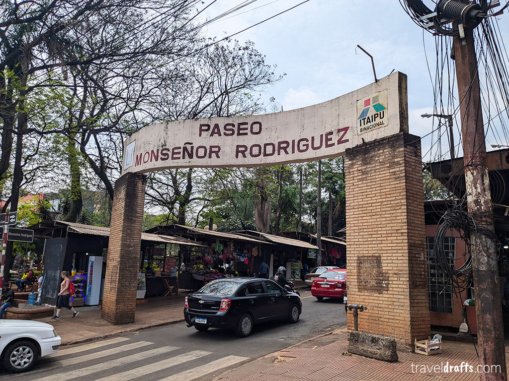 A brick archway bearing the inscription "Paseo Monseñor Rodriguez" towers over a street in Paraguay. Market stalls selling clothing and other goods line the sidewalk beneath the trees—an ideal spot for those seeking travel tips and planning to visit Paraguay.