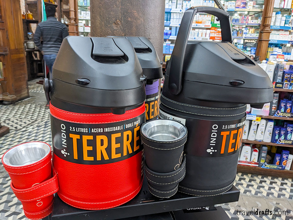 Two large thermoses labeled "TERERÉ" in red and black boxes sit on a counter with matching metal cups, highlighting a Paraguayan tradition. Pharmacy shelves filled with products can be seen in the background.