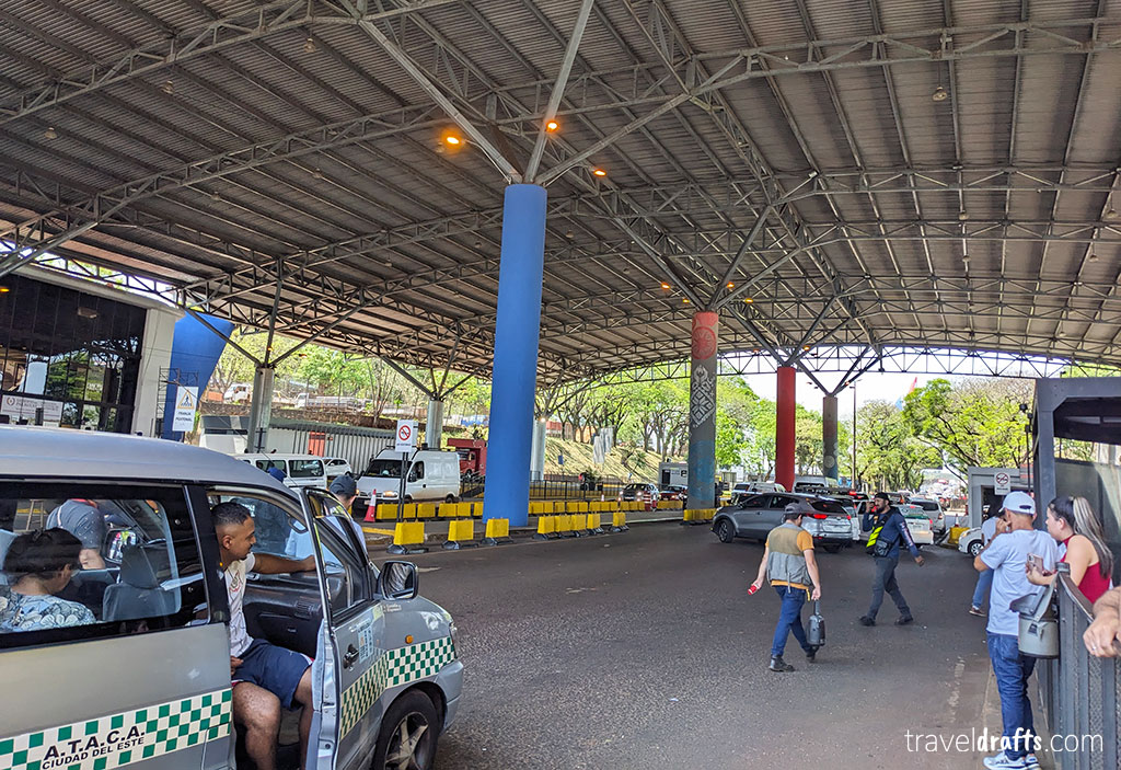A covered border checkpoint with people walking, parked cars, security personnel, and concrete pillars painted blue and red. Trees and traffic barriers are visible in the background—an essential stop for anyone visiting Paraguay or seeking travel tips.