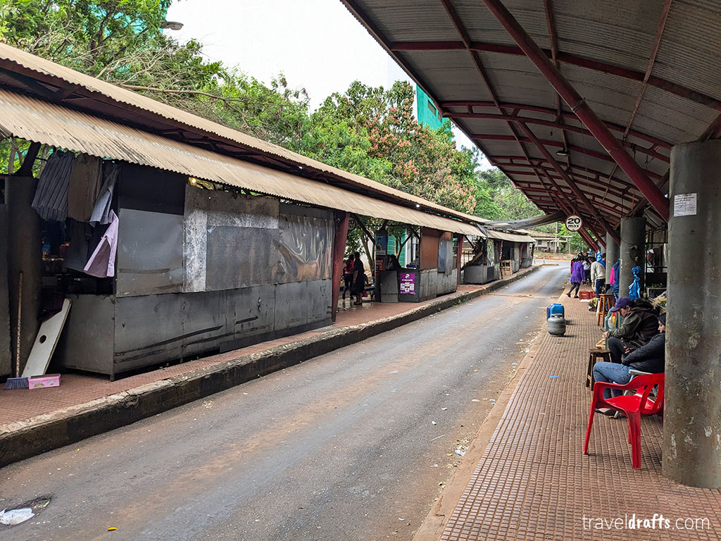 A quiet street market, with empty vendor stalls on the left and a covered walkway on the right where a few people sit on red chairs—perfect for those looking for hints of Paraguay, while trees and greenery fill the background.
