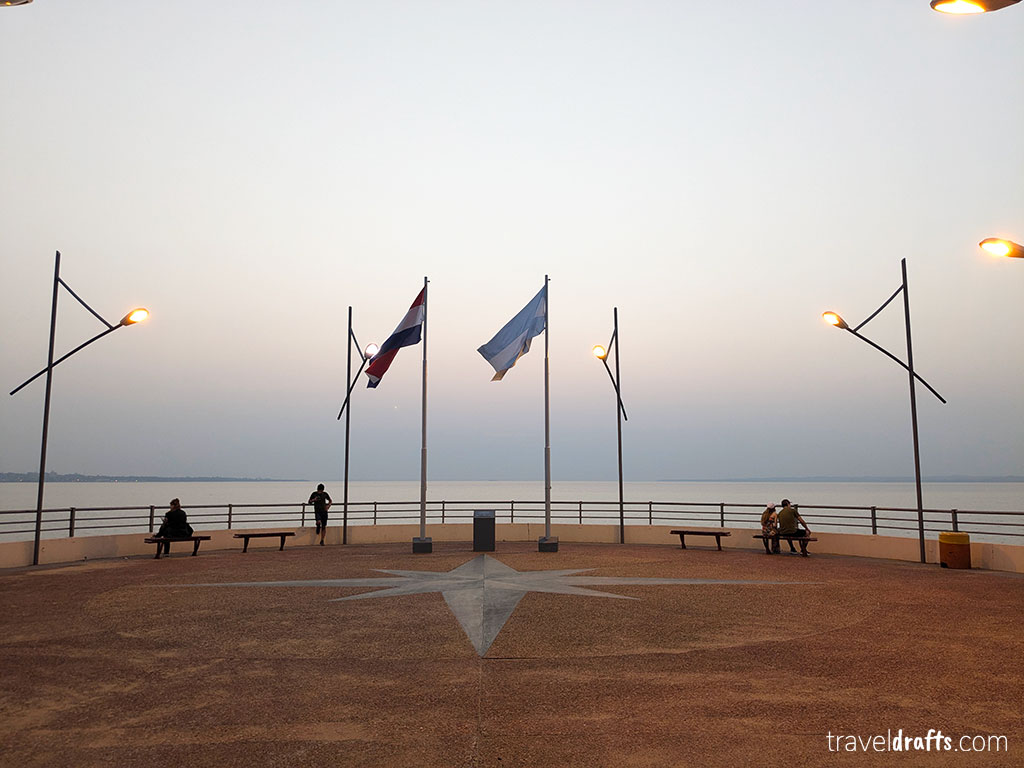Um calçadão à beira-mar no Paraguai com três mastros - duas bandeiras visíveis - bancos, postes de iluminação pública e algumas pessoas relaxando. O céu claro e o sol nascente ou poente o tornam um local perfeito para quem procura dicas de viagem antes de visitar o Paraguai.