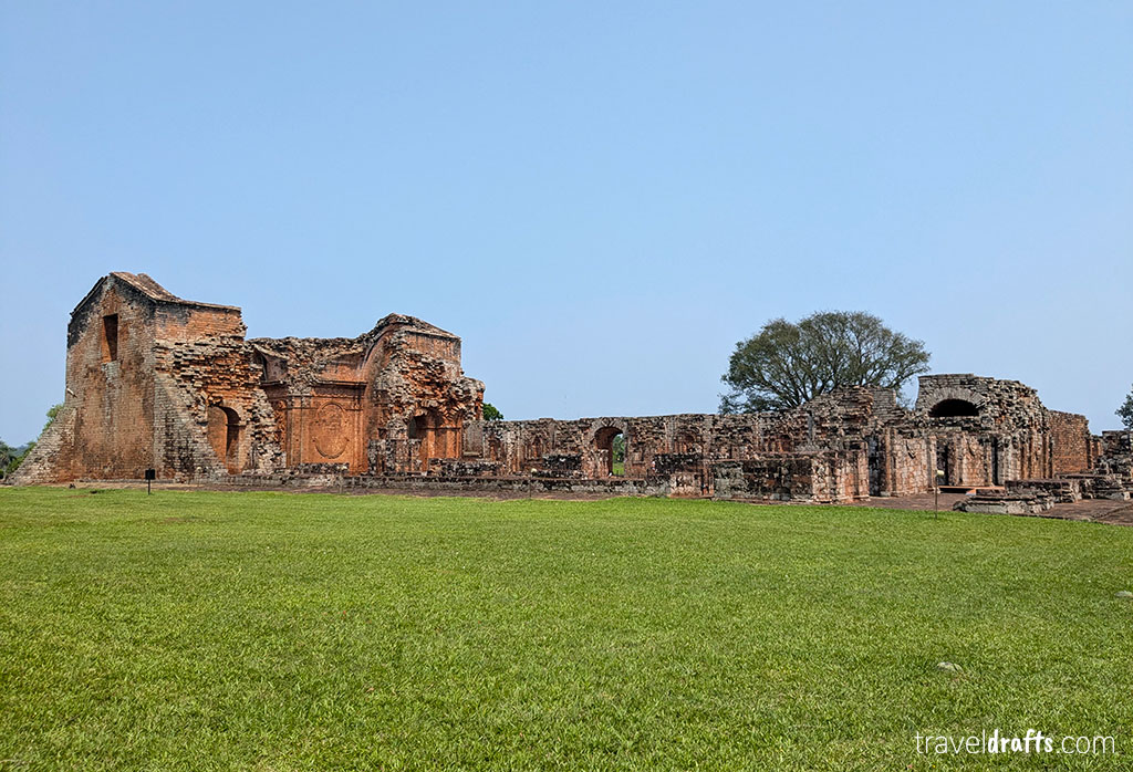 Ruins of a large stone building with red brick walls and arches stand on a grassy area under a clear blue sky. Some walls are partially collapsed, and trees are visible in the background—an ideal location for those interested in exploring Paraguay's history.