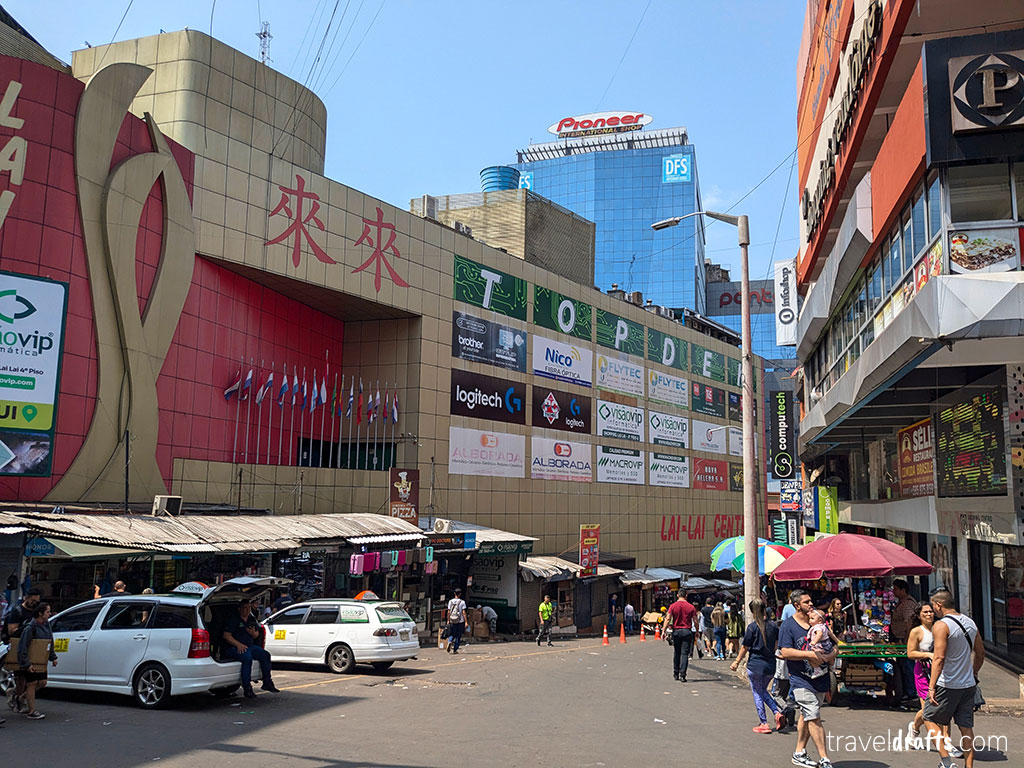 A bustling street scene featuring a modern shopping mall with signs for various brands, street vendors under colorful umbrellas, parked cars, and people walking by—perfect for anyone visiting Paraguay on a sunny day.