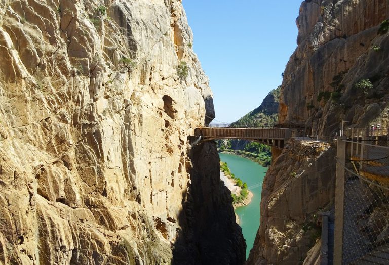 A narrow bridge passes between two passes of rocky cliffs above a river, with a plank walk and a safety tackle visible; the water below appears green under a clear blue sky.