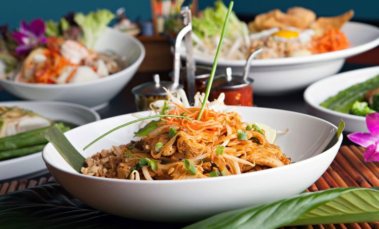 A white bowl filled with Pad Thai, Thailand's most beloved noodle dish, sits on a table topped with bean sprouts, sliced ​​carrots, green onions, chicken, and decorated with flowers—ready to reveal its secret blend of flavors.