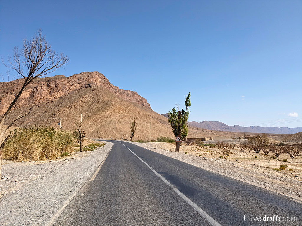 Uma estrada pavimentada curva-se através de uma paisagem desértica e seca, com vegetação esparsa e uma colina rochosa ao fundo, sob um céu azul claro, evocando a beleza cénica encontrada num passeio ao deserto do Saara.