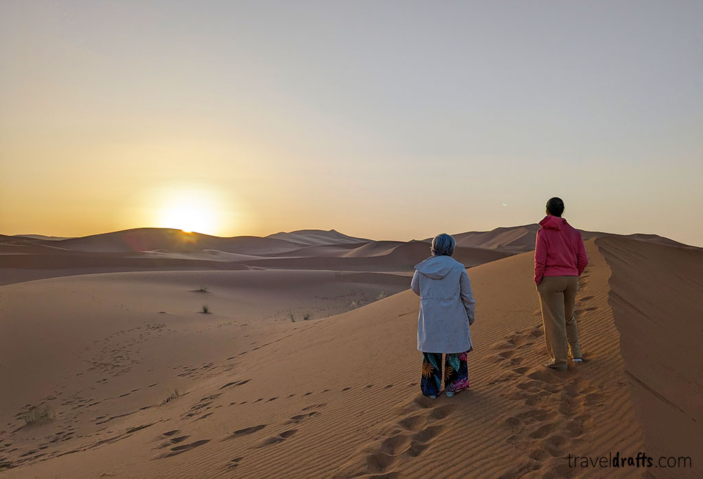 Duas pessoas estão numa duna de areia no deserto ao pôr do sol, com pegadas atrás. Uma delas usa um lenço na cabeça e um casaco comprido, a outra um casaco com capuz - captando a magia de um passeio ao deserto do Saara enquanto o sol se põe no horizonte claro.
