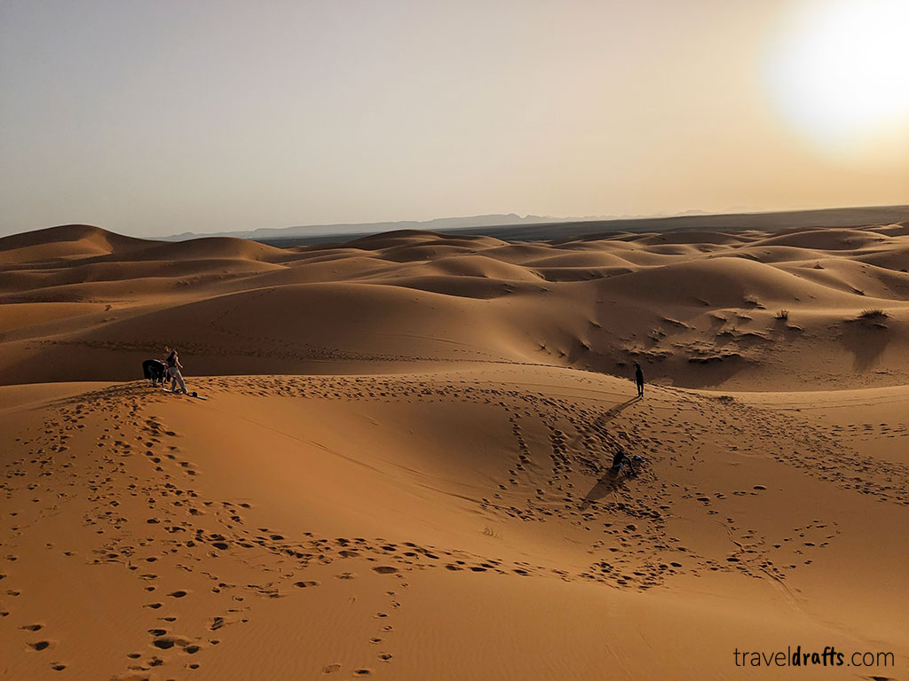 Uma vasta paisagem desértica com dunas de areia ondulantes ao pôr do sol, vários conjuntos de pegadas na areia e algumas pessoas a desfrutar de um passeio ao deserto do Saara enquanto caminham e se sentam nas dunas sob um céu limpo.