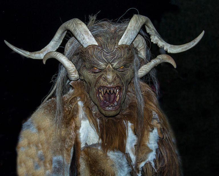 A person wears a detailed Krampus costume, with long twisted horns, sharp fangs, pointed ears, and fur-covered shoulders, set against a dark background — a striking reference to unique Christmas traditions from around the world.