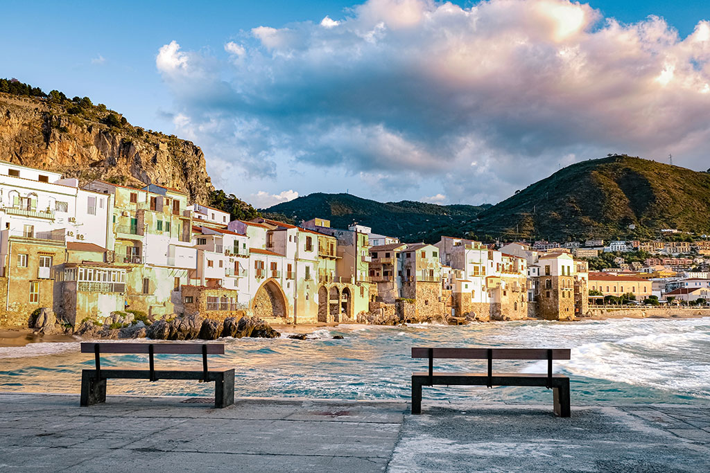 Two empty benches face the colourful historic buildings along the seafront of Cefalù, with waves crashing below and green hills rising in the background beneath a partly cloudy sky — perfect for embracing the island lifestyle and the charm of local travel.