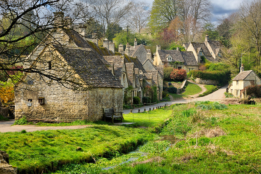 A row of old stone houses with steep roofs and tiles lines a winding village road — perfect for the peaceful getaways UK travellers look for. Lush grass, shrubs, and a small stream run alongside, framed by trees beneath a cloudy sky.