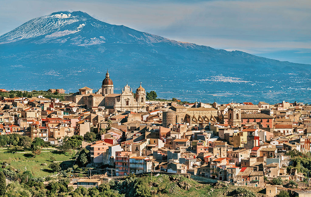 In the foreground, a picturesque local town with historic buildings and churches, while Mount Etna dominates the landscape of the beautiful island of Sicily beneath a blue sky.