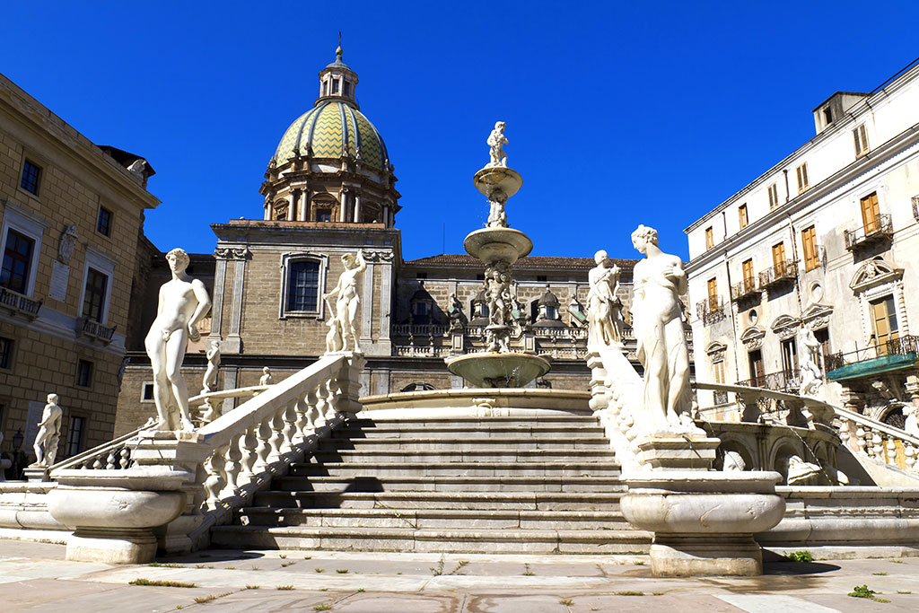 An ornate stone fountain with several statues and staircases stands in front of historic island buildings and a domed church beneath a clear blue sky in Sicily.