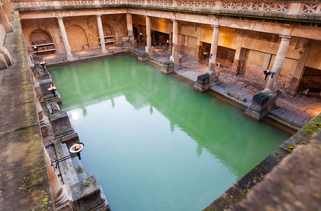 A view of the historic Roman Baths in Bath — an ideal destination for peaceful getaways — shows a large rectangular pool surrounded by ancient columns and stone walls, with greenish water reflecting the timeless architecture.
