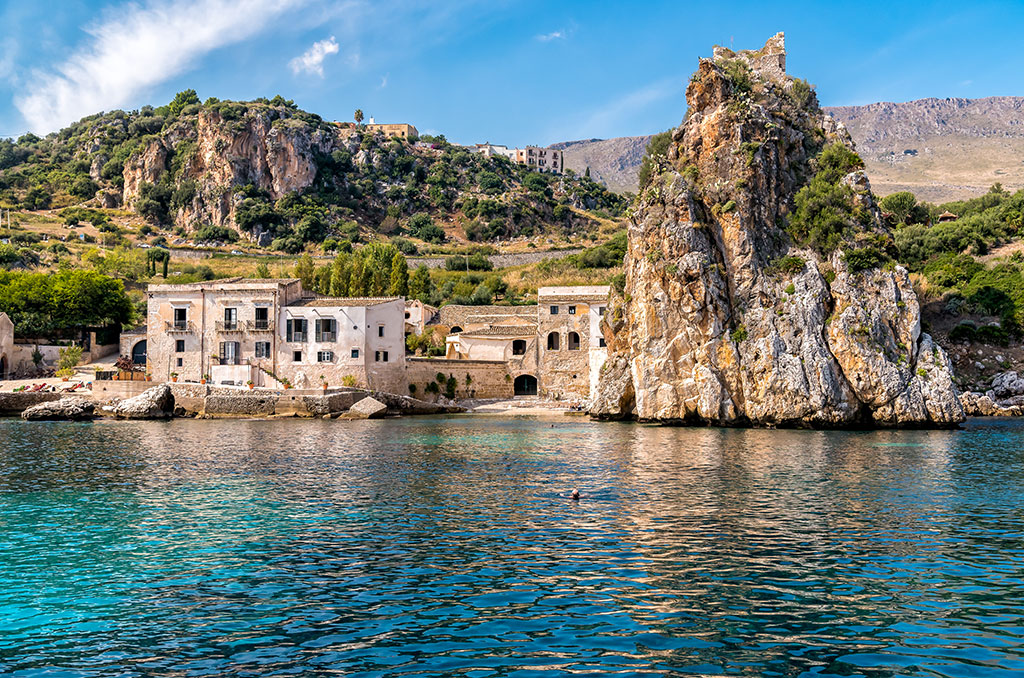 A coastal scene in Sicily, with old stone buildings by the water, a rocky outcrop beside them, green hills beyond, and a blue sky overhead. The calm sea in the foreground reflects the landscape and captures the island’s relaxed pace of life.