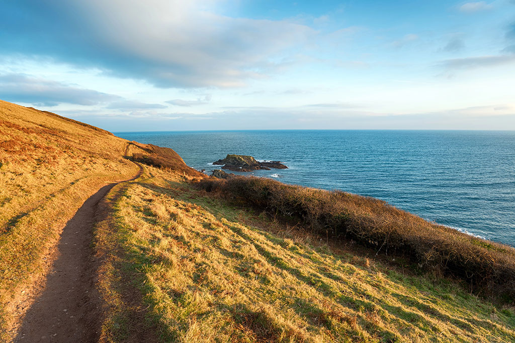 A dirt path winds along a grassy hillside overlooking the ocean, with a rocky outcrop visible in the water — an idyllic setting perfect for peaceful retreats in the UK. The sky is partly cloudy, with sunlight illuminating the landscape.